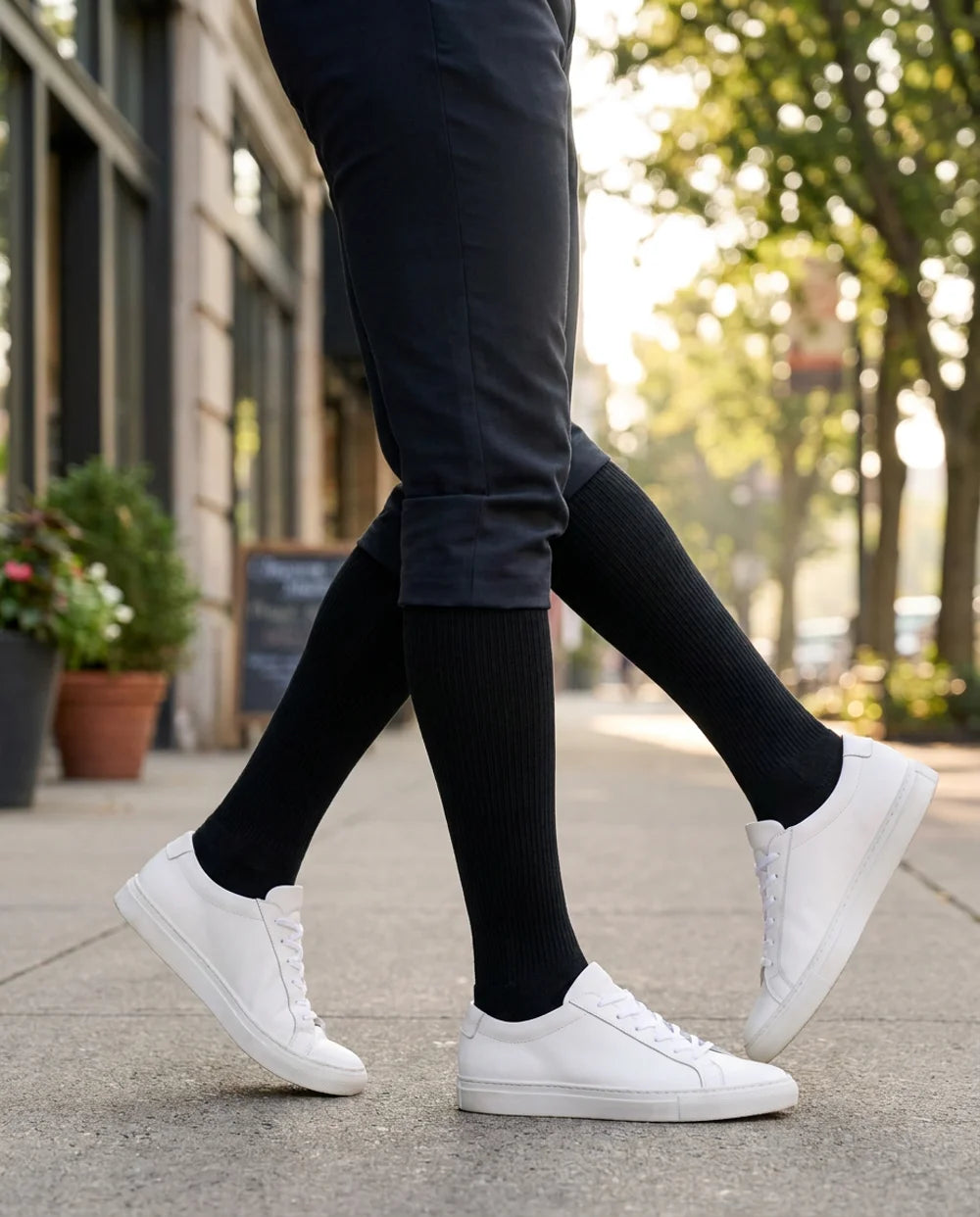 Mid-stride on a tree-lined sidewalk, black Ovena knee-high compression socks into white sneakers